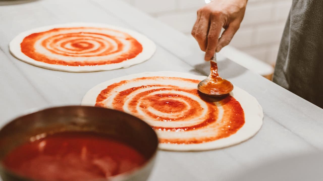 Person spreading tomato sauce on pizza dough with a spoon, two pizza bases on a white surface, metal bowl filled with tomato sauce, preparation in progress, indoor setting