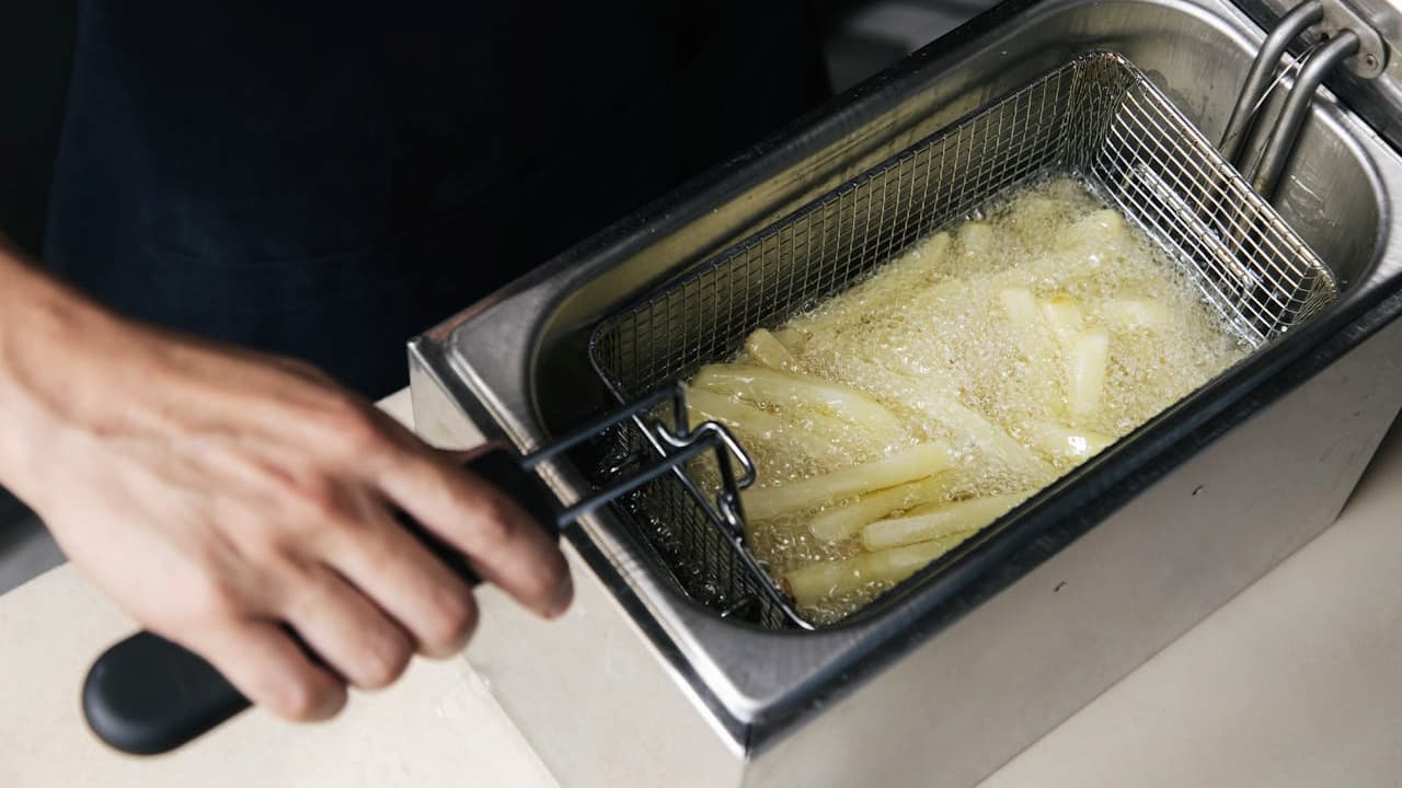 Person preparing food, hand holding basket of raw French fries, fries placed in deep fryer, stainless steel fryer with oil, kitchen countertop visible, cooking in progress, food preparation scene, fast food or home kitchen setting