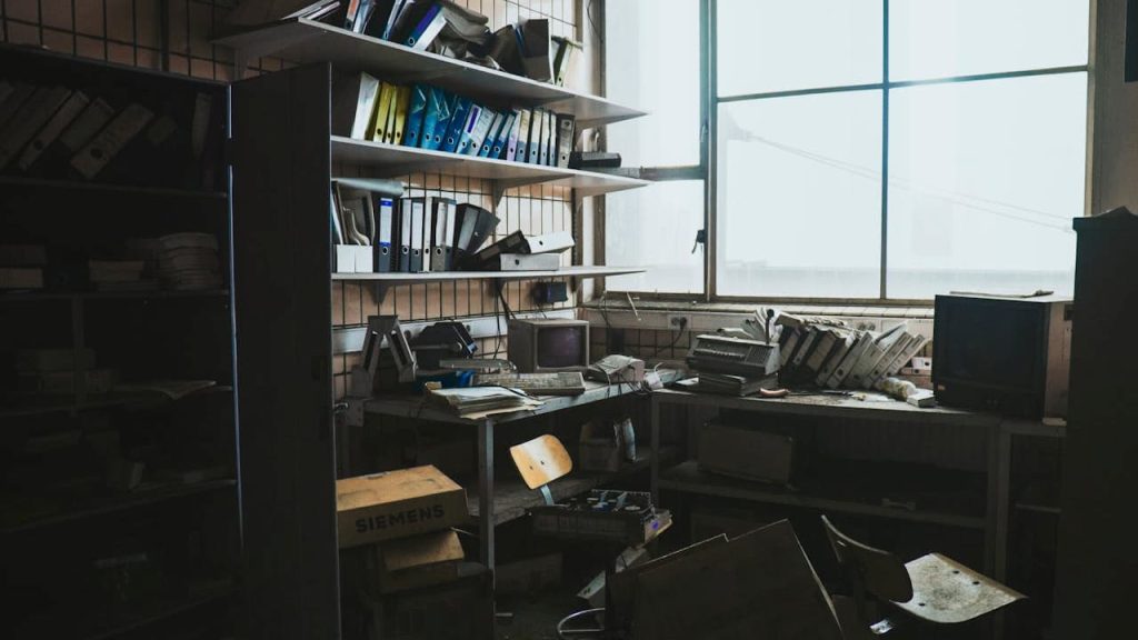 Clutter covering window, stacked boxes, plastic bins, old clothes, papers, and miscellaneous household items blocking natural light, dusty glass panes, partially drawn curtains