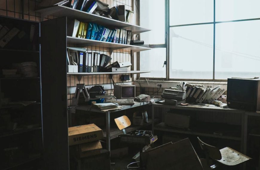 Clutter covering window, stacked boxes, plastic bins, old clothes, papers, and miscellaneous household items blocking natural light, dusty glass panes, partially drawn curtains