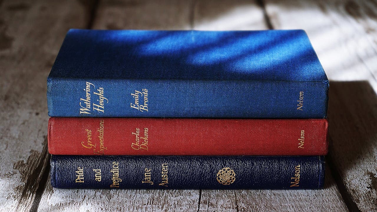 Three hardcover books stacked on a wooden surface, top book is blue with Chinese characters and the number 1 on the spine, middle book is red with Chinese characters and the number 2, bottom book is dark blue with gold Chinese characters and the number 3, light and shadow patterns fall across the books