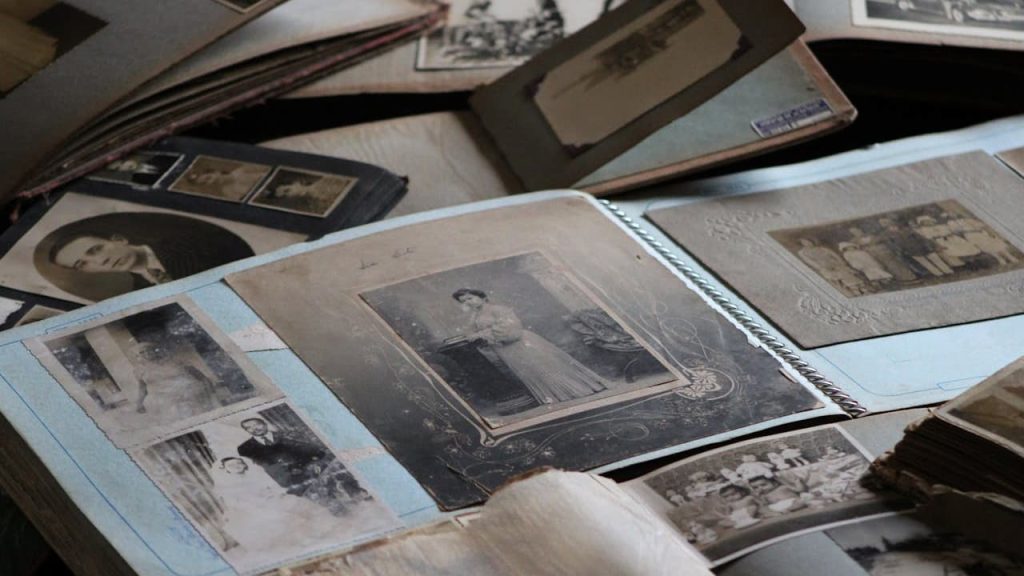 Open vintage photo albums spread out on a table, sepia and black-and-white portraits mounted on blue pages, handwritten notes beside some photos, delicate preservation materials nearby, nostalgic atmosphere of memory keeping