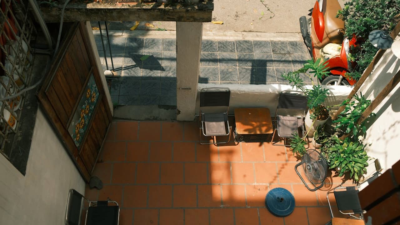 Small sunlit patio, terracotta tile flooring, wooden table and chairs, potted plants along the wall, metal railing casting shadows, street view in background, cozy and inviting outdoor space