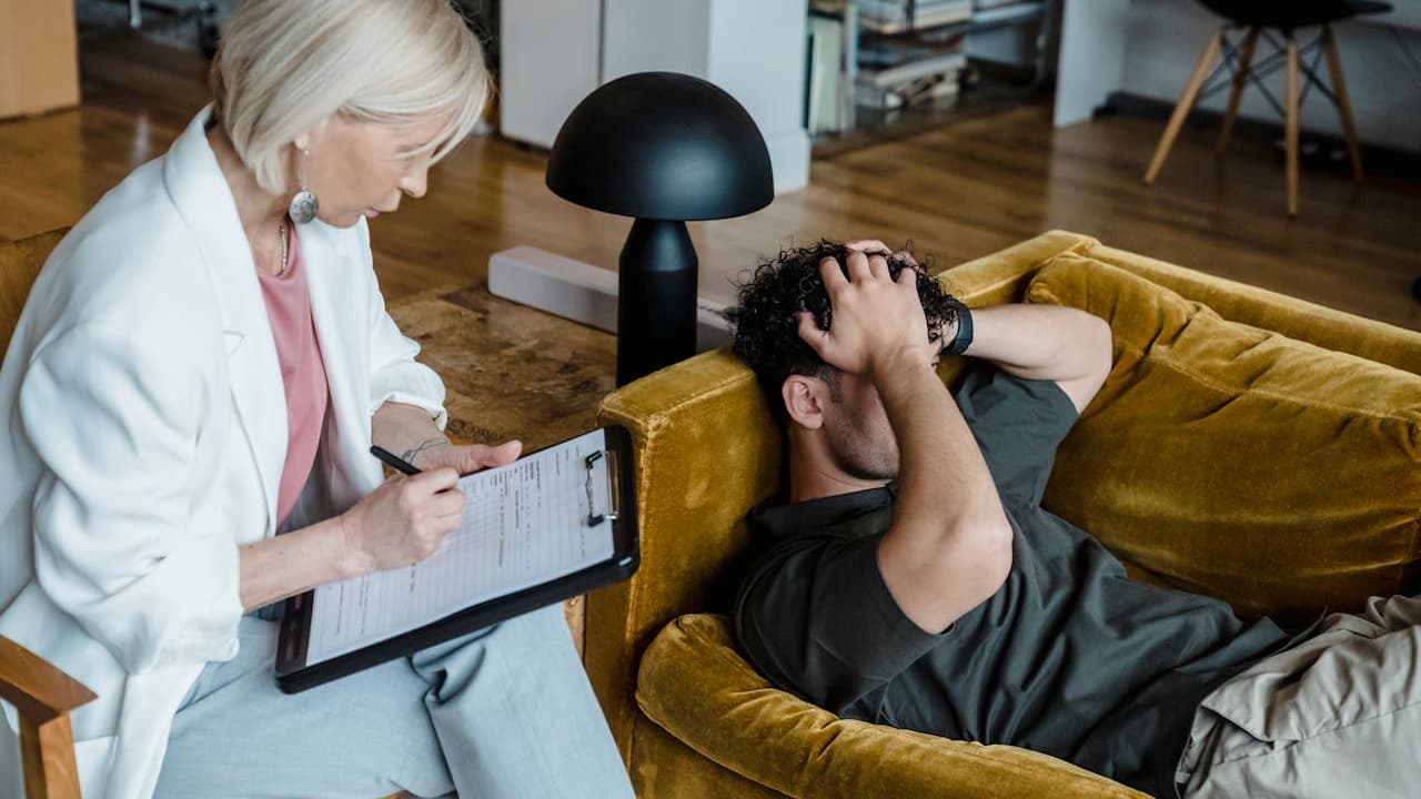 Older female therapist with short white hair, wearing a white blazer, writing on a clipboard, sitting beside a distressed man lying on a mustard yellow couch, hands covering his face, in a modern, warmly lit room