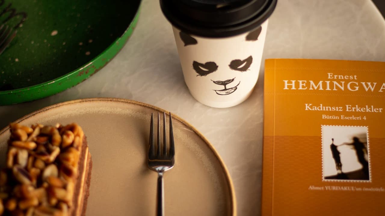 Plate with waffle and fork, panda-themed coffee cup, orange book titled “The Essential Hemingway” by Ernest Hemingway, all placed on a light-colored surface