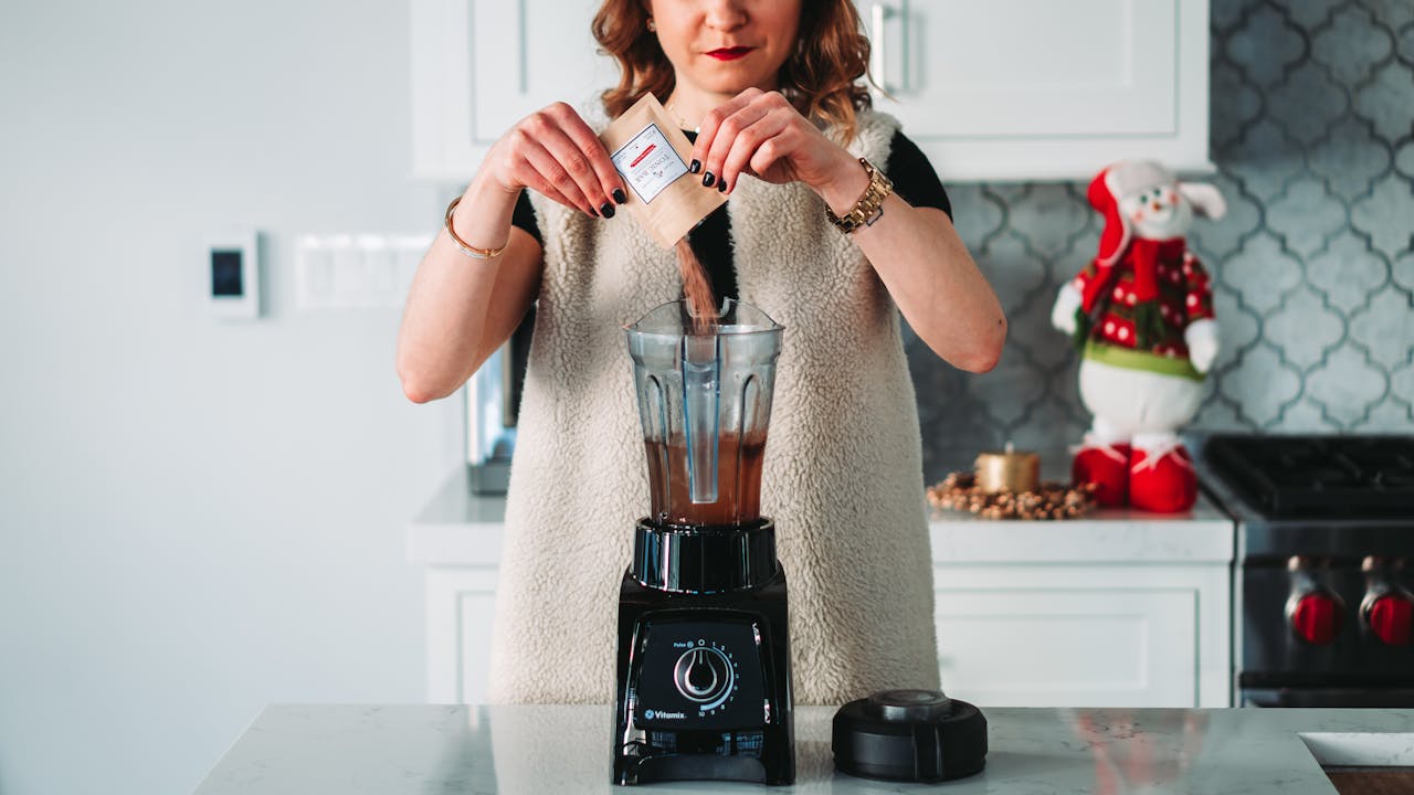 Woman pouring powder into blender with chocolate mixture, standing in kitchen, snowman decoration in background