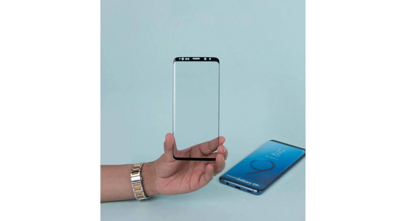 Person holding transparent smartphone screen protector, Samsung Galaxy S9 lying on flat surface in background