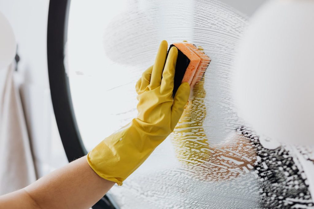 Hand in yellow glove scrubbing soapy glass surface with sponge, circular mirror in background