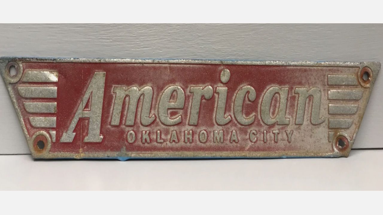 Vintage metal sign with embossed text “American Oklahoma City,” red background, silver lettering, four corner screw holes, aged and slightly rusted
