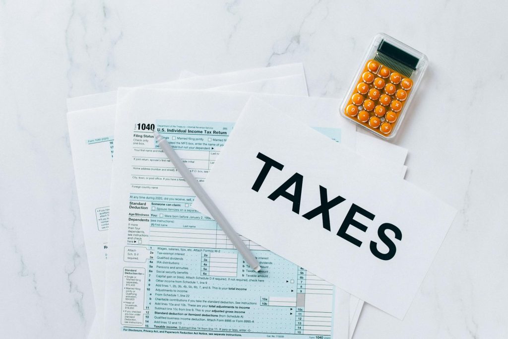 Tax forms on a white surface, paper labeled "TAXES," silver pen resting on the forms, orange calculator nearby, top-down view