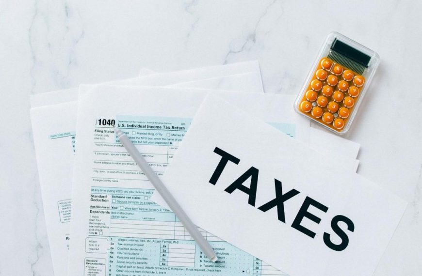 Tax forms on a white surface, paper labeled "TAXES," silver pen resting on the forms, orange calculator nearby, top-down view