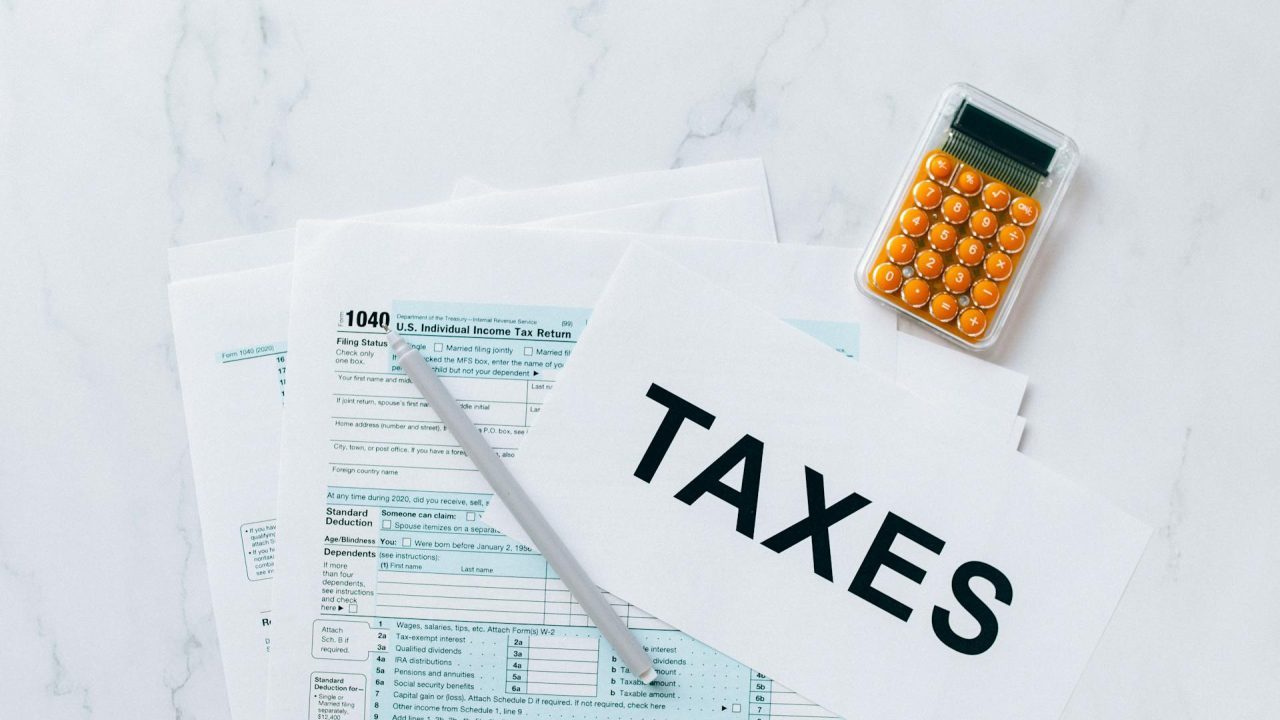 Tax forms on a white surface, paper labeled "TAXES," silver pen resting on the forms, orange calculator nearby, top-down view