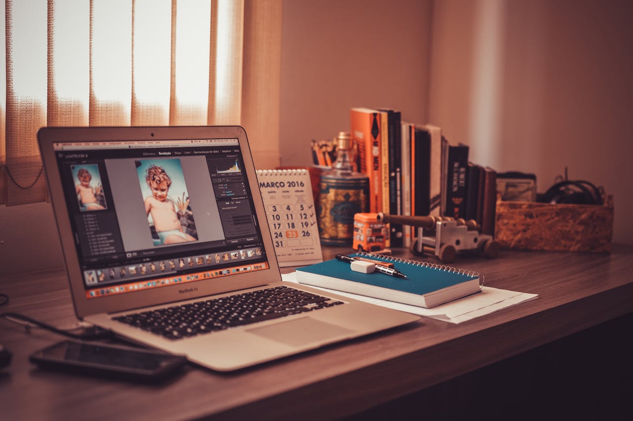  Laptop displaying photo editing software on a desk with a calendar, books, notebook, and decorative items