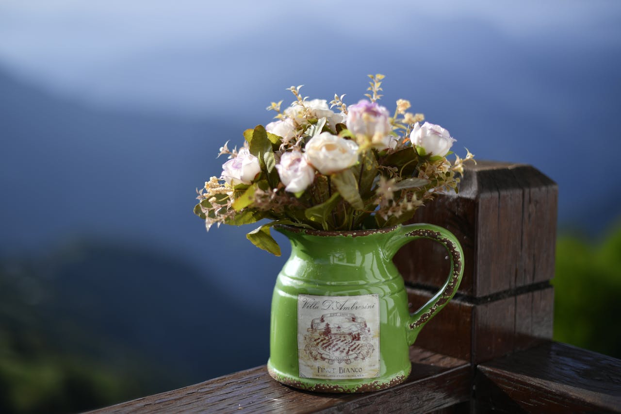 Vintage green ceramic pitcher used as a flower vase, placed on a wooden railing with mountain view backdrop