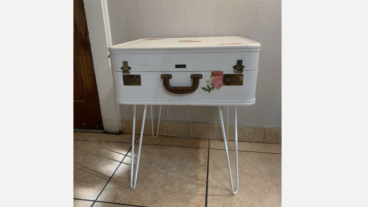 vintage white suitcase, brown handle, floral decal, mounted on white metal hairpin legs, used as bedside table, placed on tiled floor, against light-colored wall