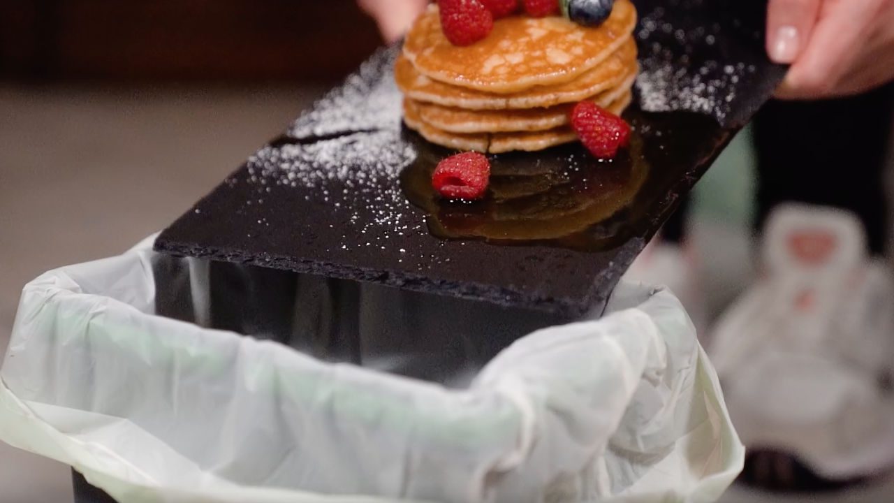 Stack of pancakes with berries and syrup being thrown into a trash can lined with a white bag