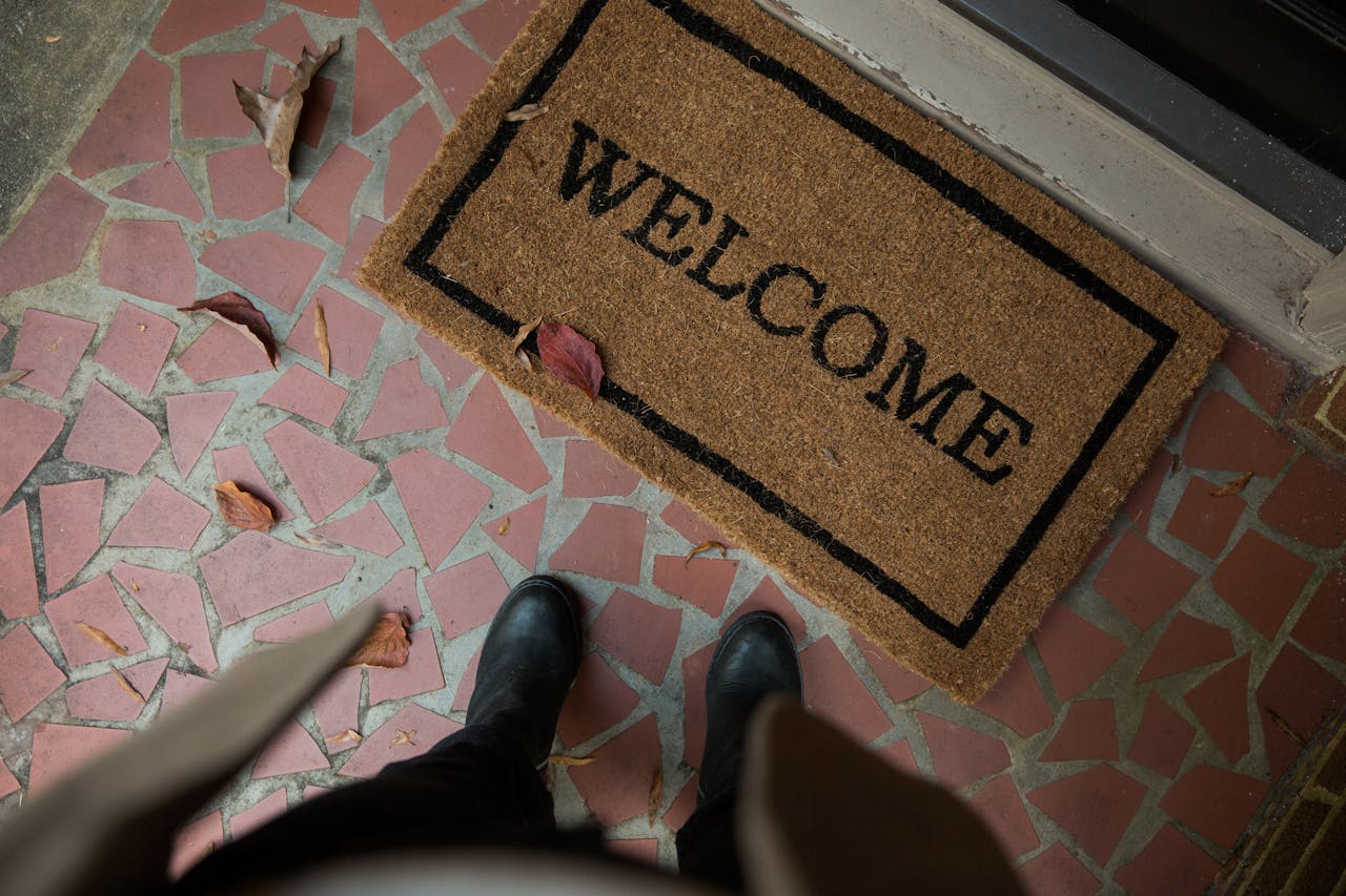 Welcome mat on a tiled doorstep with scattered leaves and a person standing in front of it