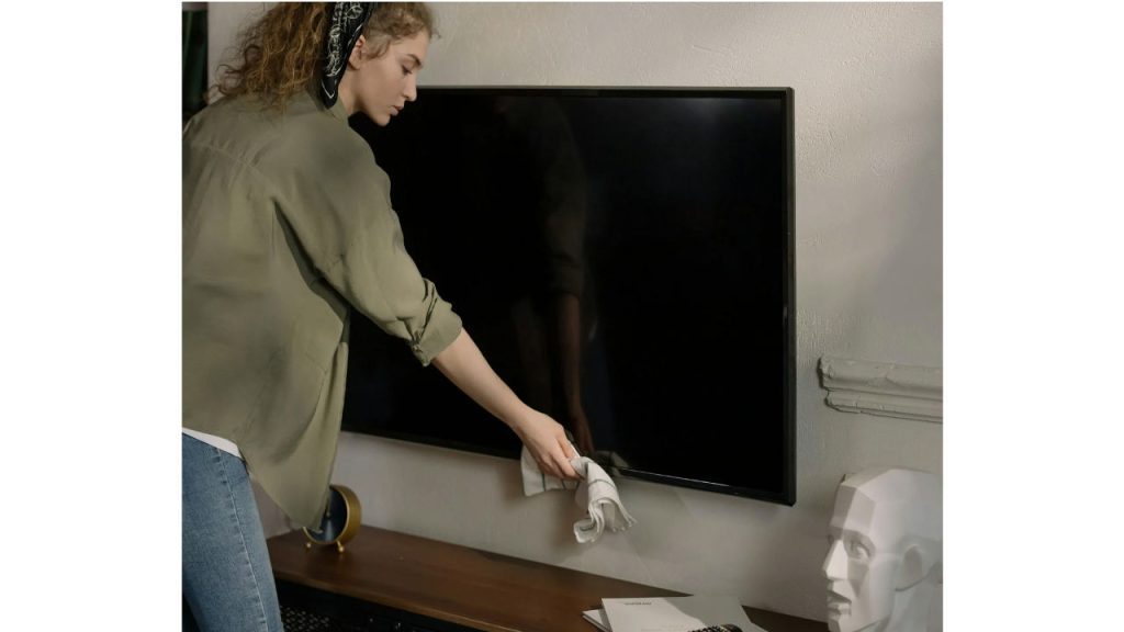 Woman cleaning flat-screen TV with cloth, standing in living room, wearing green shirt and headband