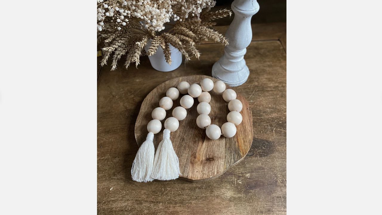 Wooden bead garland with white tassels on a round wooden tray beside dried flowers and a white candlestick