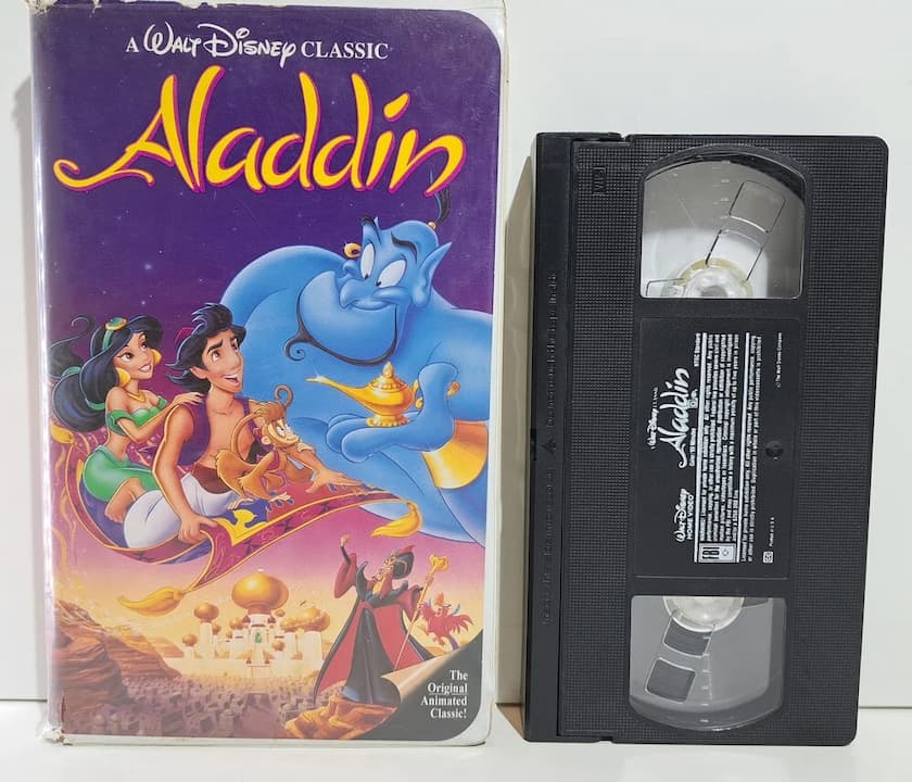 Disney’s Aladdin Black Diamond VHS edition, clamshell case with colorful artwork featuring Aladdin, Jasmine on the magic carpet, Genie hovering above them, ornate golden palace in the background, spine labeled “Walt Disney Classic,” VHS tape labeled “Aladdin,” placed on a surface, likely part of a vintage Disney collection