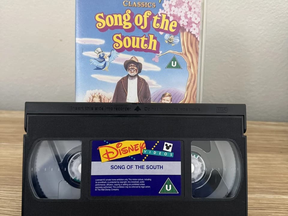 Disney VHS tape and case for Song of the South, cover shows animated birds and a smiling man in a hat with a boy behind him, label on VHS reads “Song of the South” with a Disney Videos logo, set on wooden surface against a white wall