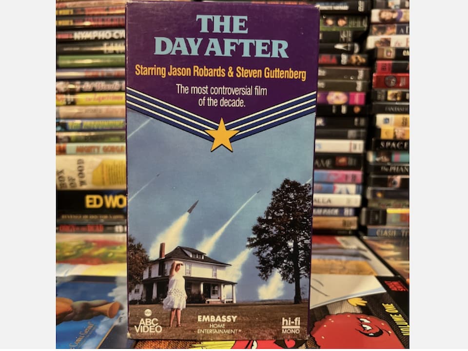 VHS tape and case for The Day After (1983), cover art showing a nuclear explosion over a city skyline, dramatic orange and black color scheme, title in bold white letters, tagline emphasizing the film’s emotional and social impact
