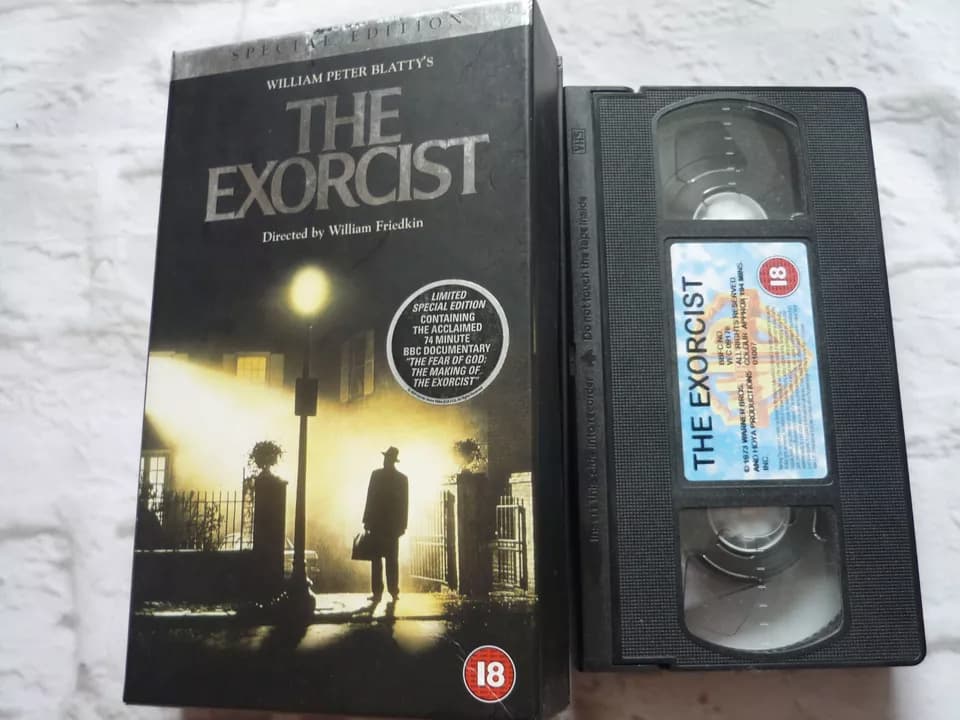The Exorcist 1981 VHS release, dark moody cover with silhouette of priest under streetlamp, Warner Home Video branding, clamshell or slipcase with classic horror typography, tape visible beside