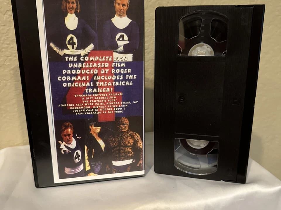 VHS tape and case of The Fantastic Four (1994) unreleased film, cover features all four main characters in blue superhero costumes, text highlights Roger Corman’s production and inclusion of original theatrical trailer, set against a beige wall and white cloth surface