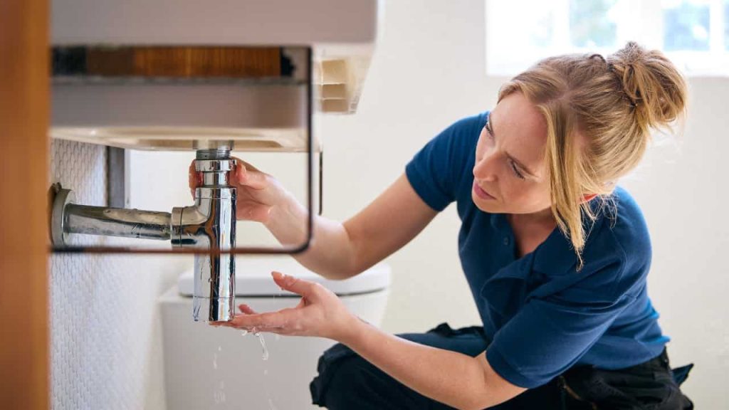 Person in blue shirt crouching near bathroom sink, examining or repairing faucet plumbing, hands positioned under sink fixture, bathroom interior with mirror visible in background
