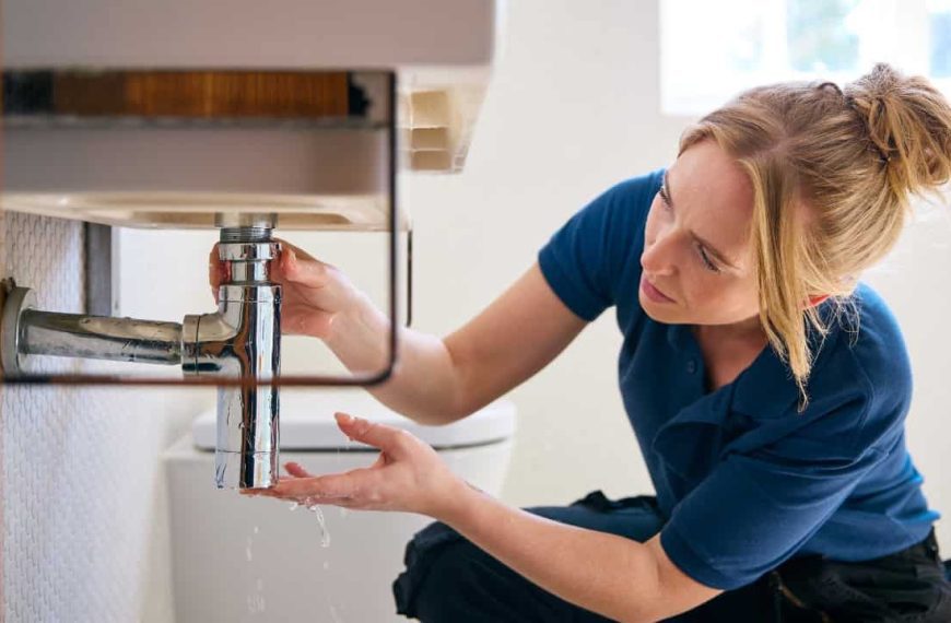 Person in blue shirt crouching near bathroom sink, examining or repairing faucet plumbing, hands positioned under sink fixture, bathroom interior with mirror visible in background