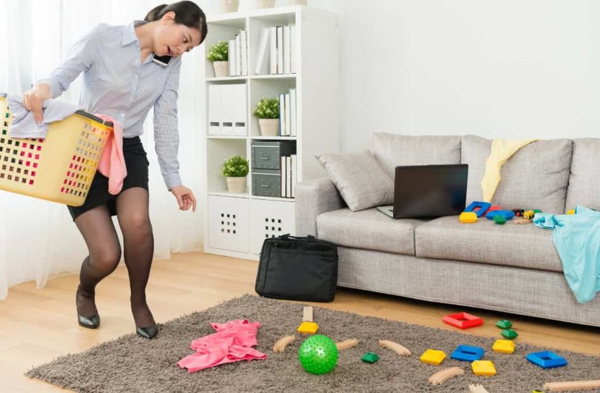 A woman in office attire, bending down to pick up toys and scattered objects, child’s play items on a rug, casual living room setting, sofa in the background