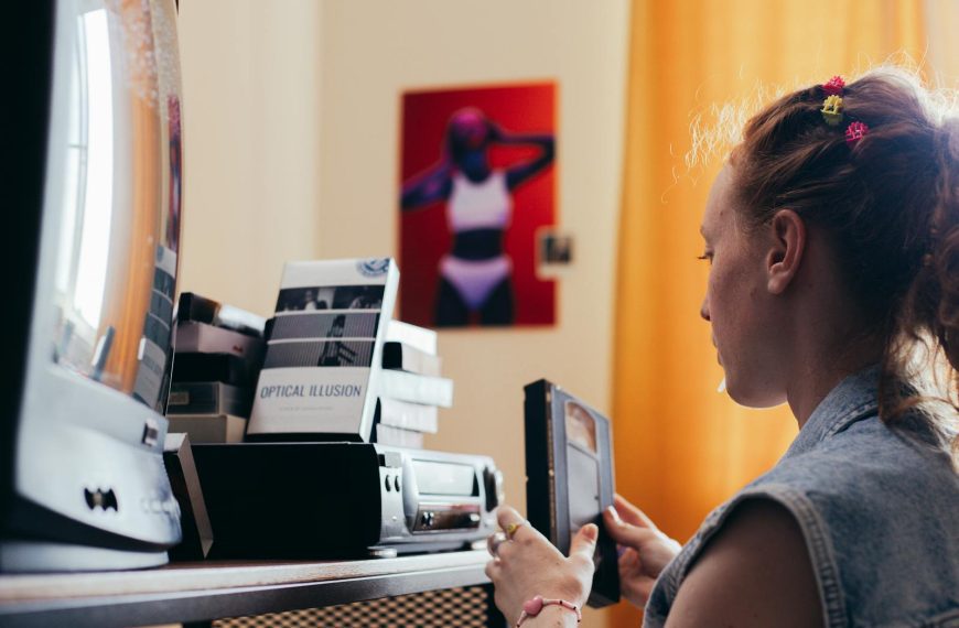 A young woman in a room holding a VHS tape, surrounded by retro equipment and decor.