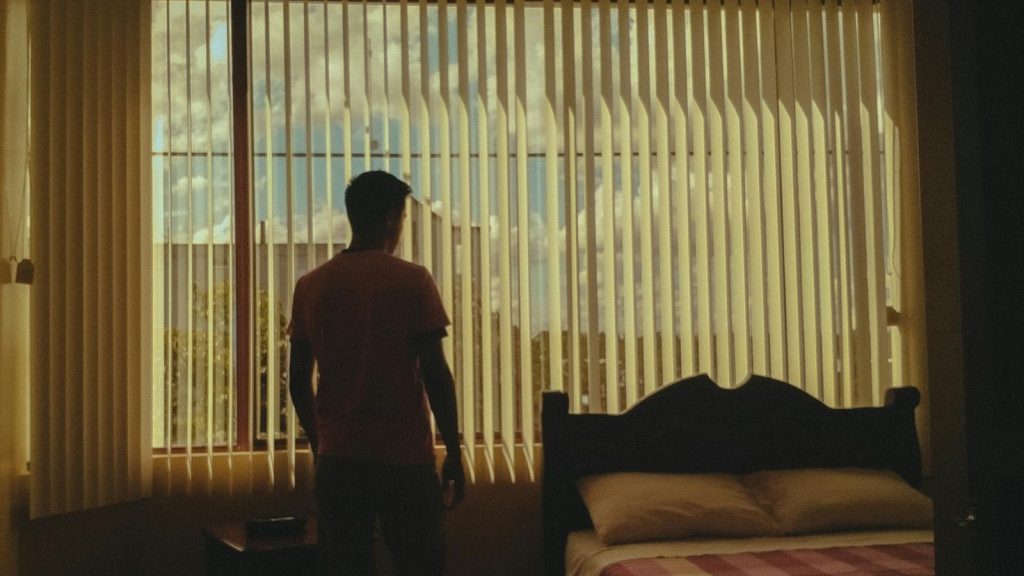 A silhouette of a man stares out a bedroom window with closed blinds, emphasizing introspection.