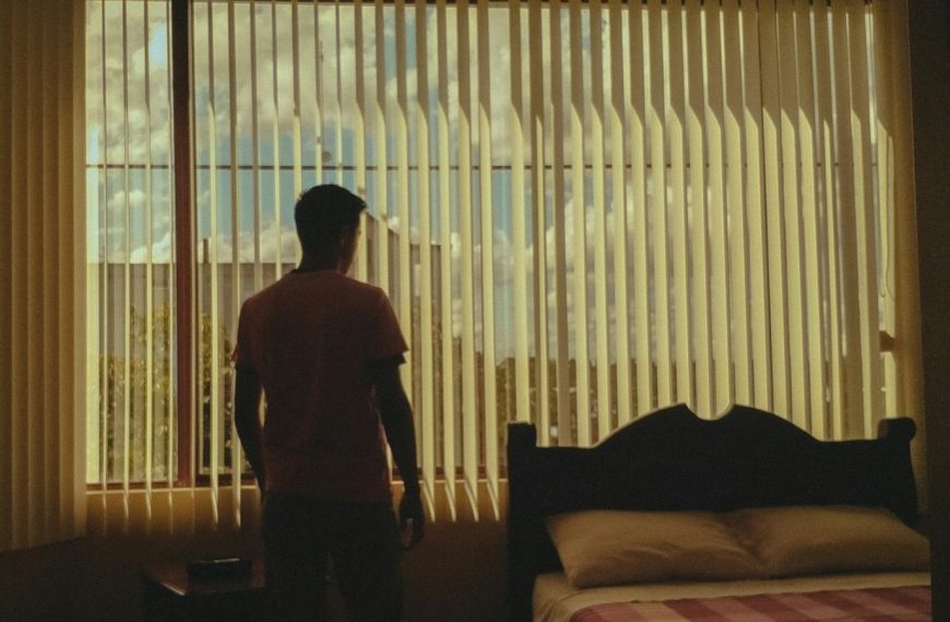 A silhouette of a man stares out a bedroom window with closed blinds, emphasizing introspection.
