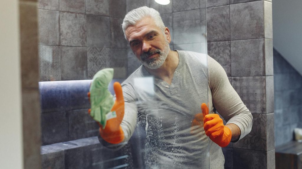Grey haired male with beard in orange rubber gloves attentively cleaning shower glass in bathroom