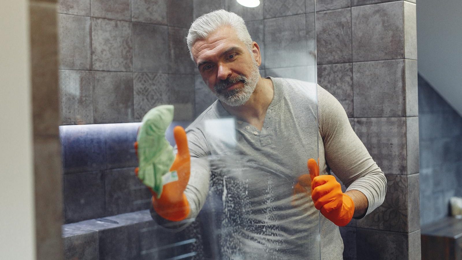 Grey haired male with beard in orange rubber gloves attentively cleaning shower glass in bathroom