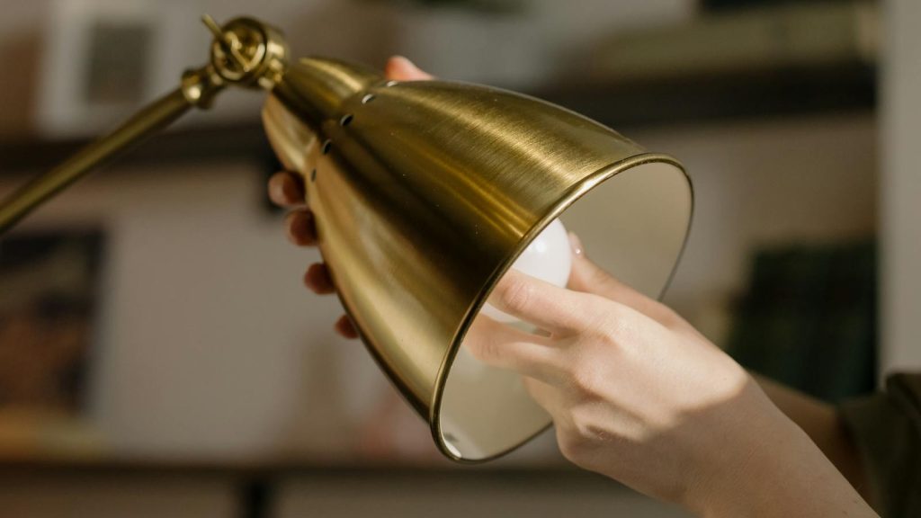 A person changing a bulb in a stylish gold desk lamp inside a cozy home office.