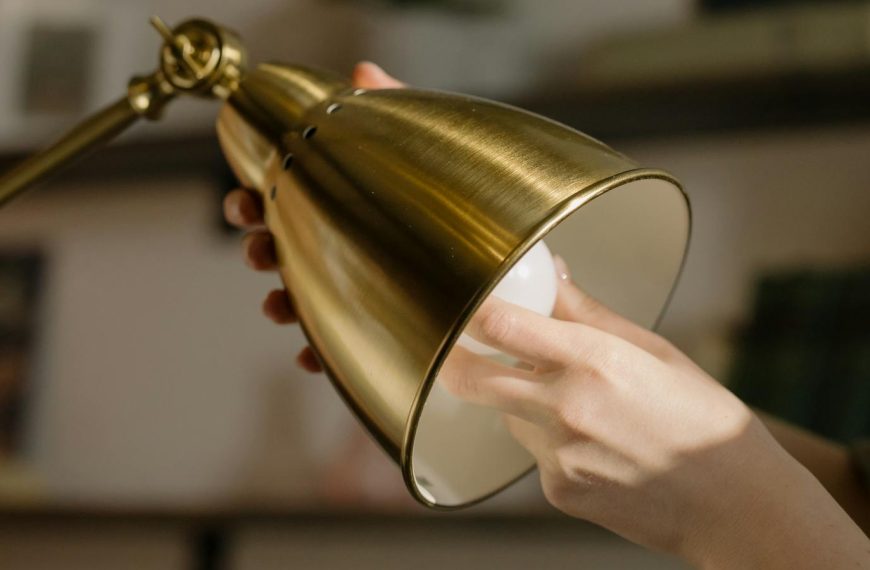 A person changing a bulb in a stylish gold desk lamp inside a cozy home office.