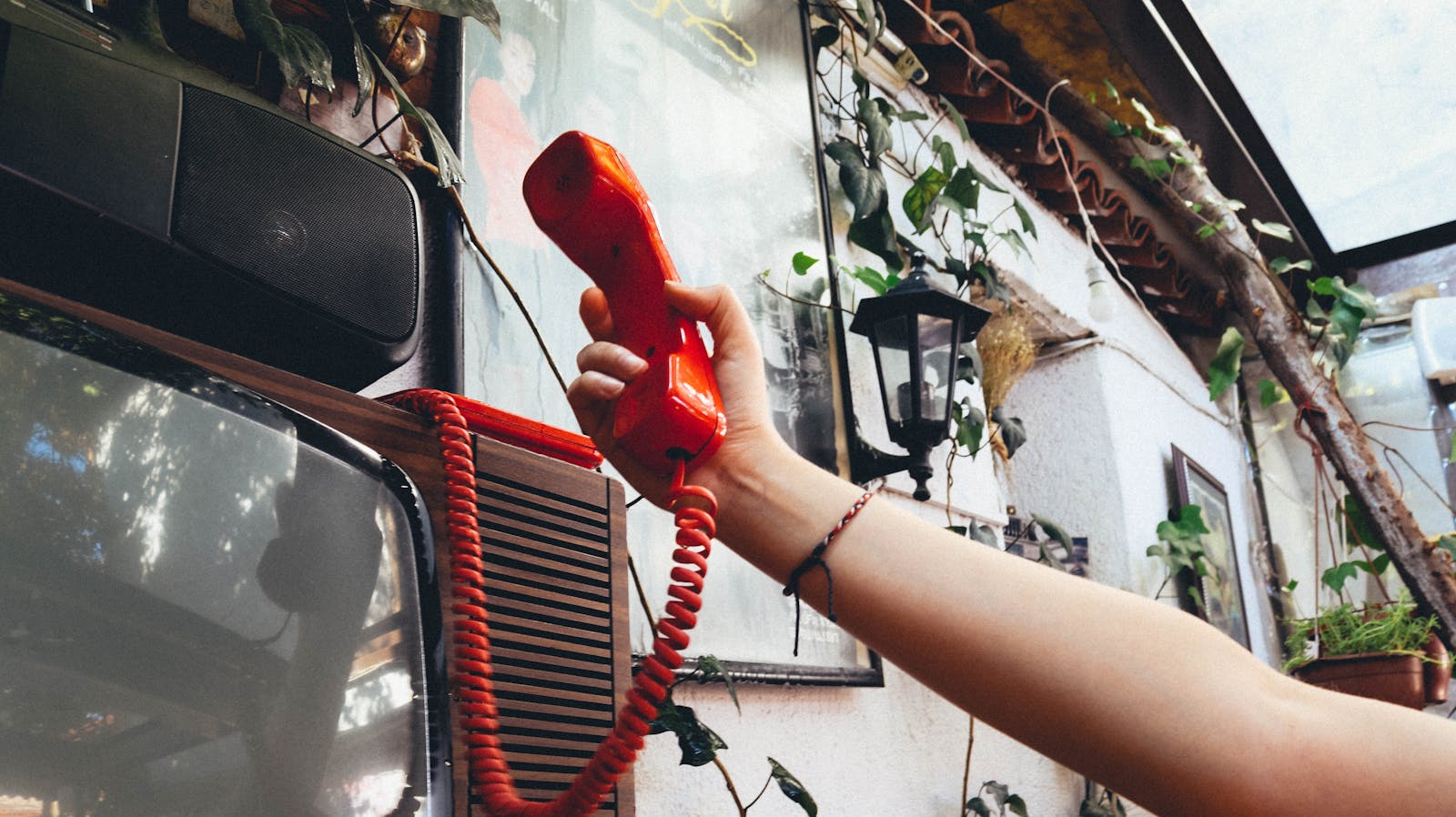 Close-up of a hand holding a nostalgic red phone in a vibrant indoor space filled with greenery.