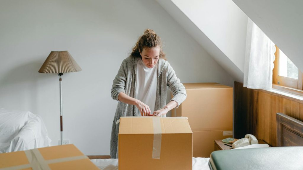 Slim female in cardigan unpacking cardboard box put on bed close to suitcase while standing near window in attic style room in daylight