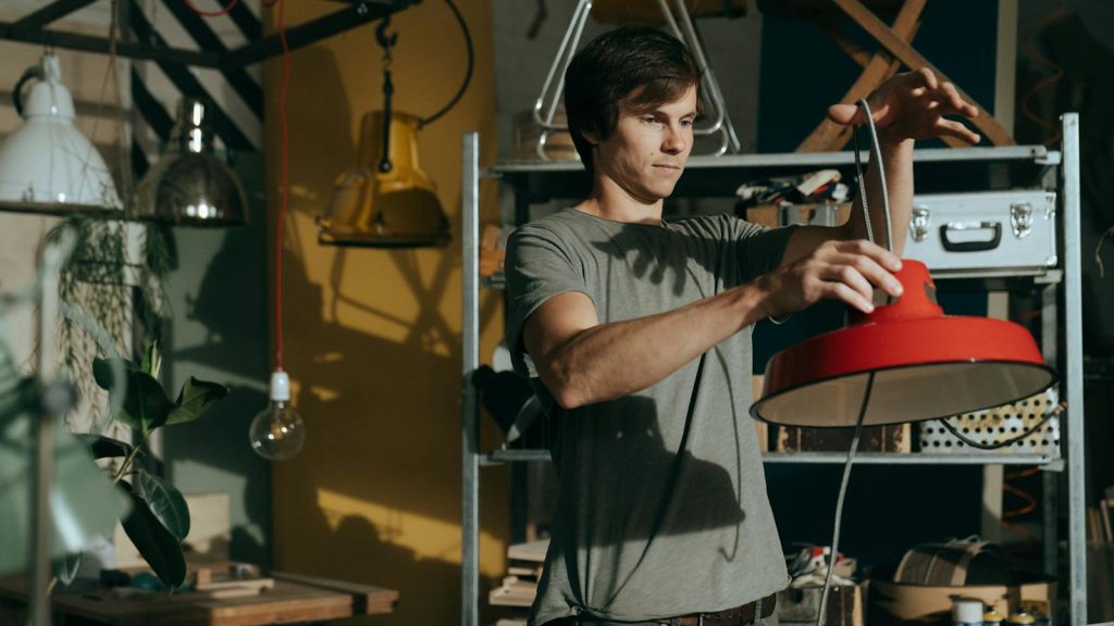 A man working indoors repairing a red lamp in a craft workshop environment.