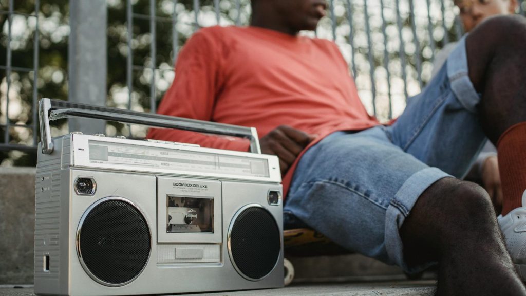 From below of crop young African American male friends listening to music on retro cassette recorder with radio while sitting near metal grid of park