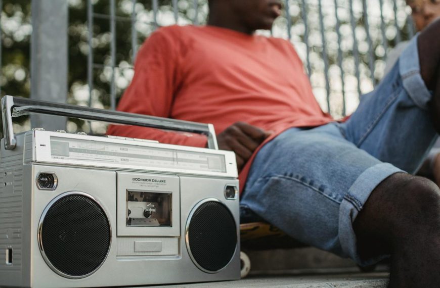 From below of crop young African American male friends listening to music on retro cassette recorder with radio while sitting near metal grid of park