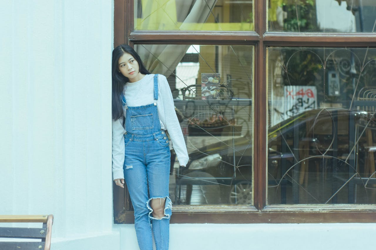 Woman in Blue Denim Jumper Standing Beside Glass Window