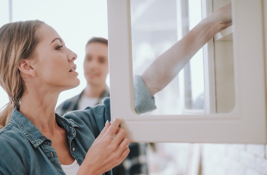 Young woman opening the glass door of the cupboard and putting her hand on the shelf. Man standing on the background