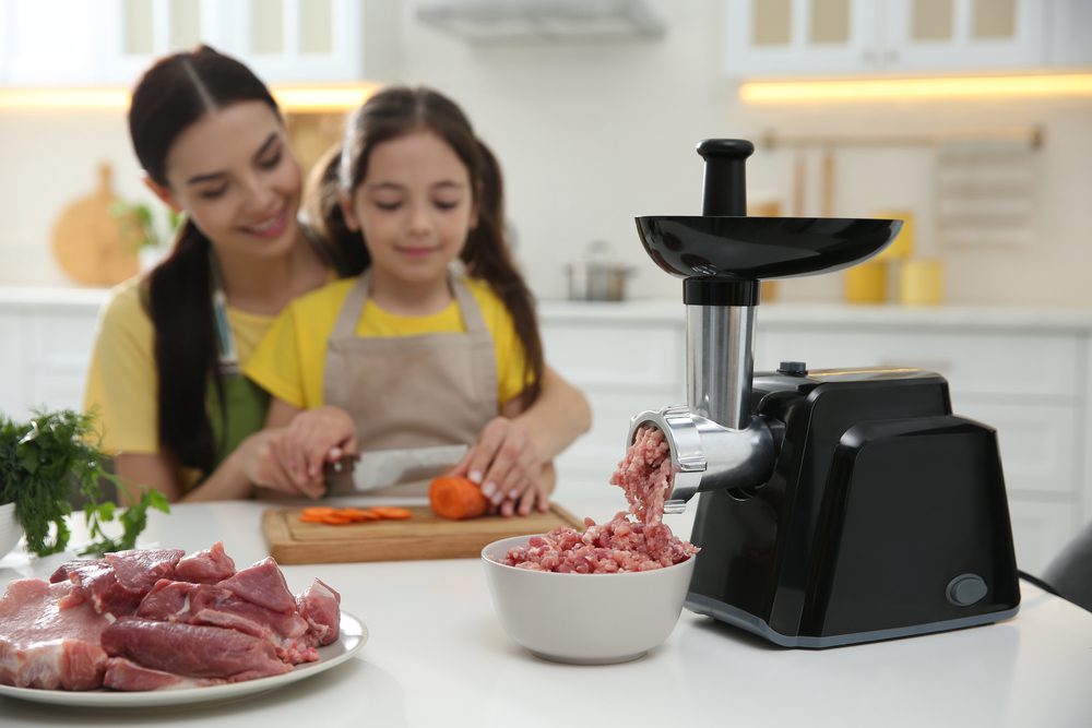 Mother and little girl cutting carrot in kitchen, focus on meat grinder