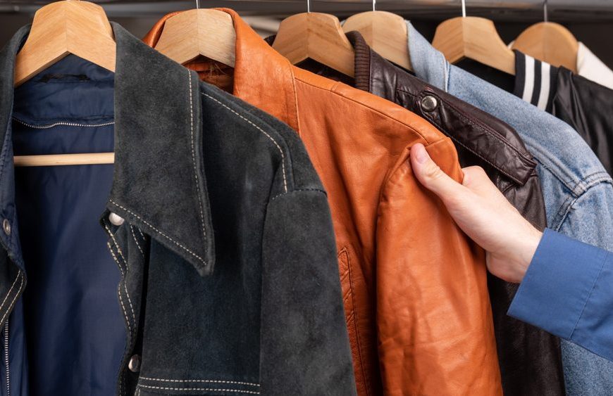 Man choosing clothing in a second hand store. Various vintage suede leather and jeans jackets hang on clothing rack. Thrifting and sustainability in clothing concept