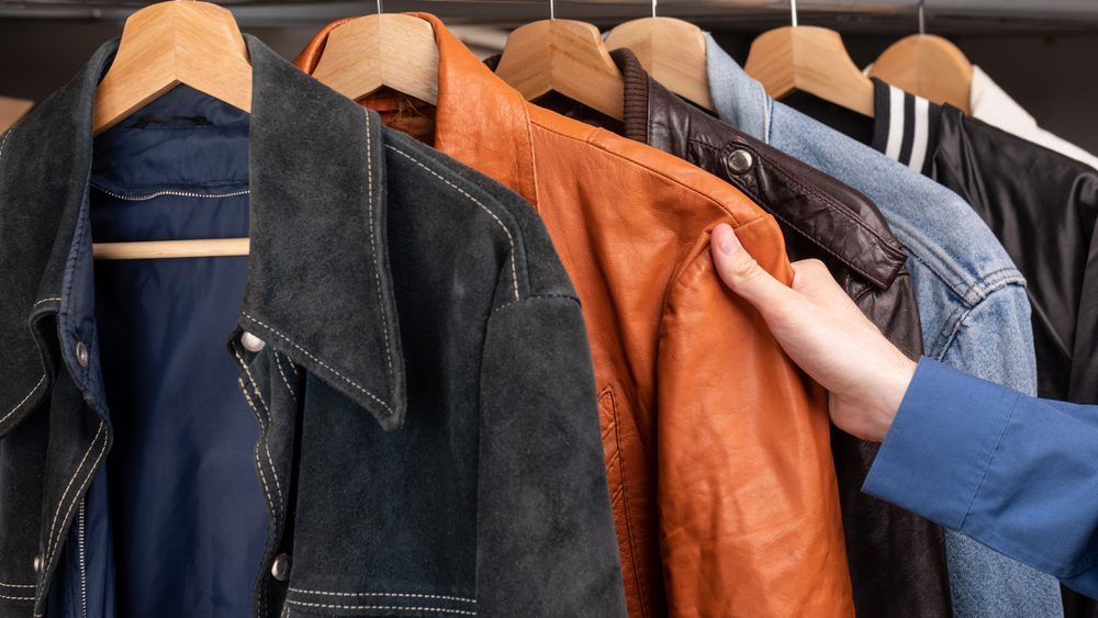 Man choosing clothing in a second hand store. Various vintage suede leather and jeans jackets hang on clothing rack. Thrifting and sustainability in clothing concept