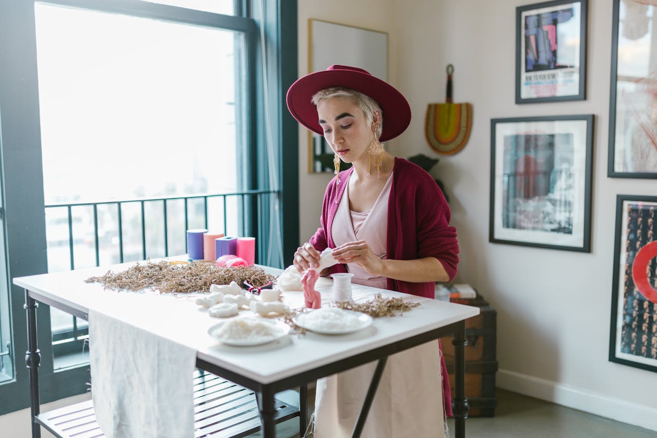 Woman in Pink Fedora Hat and Cardigan Sweater Standing In Front of Arts and Crafts on the Table