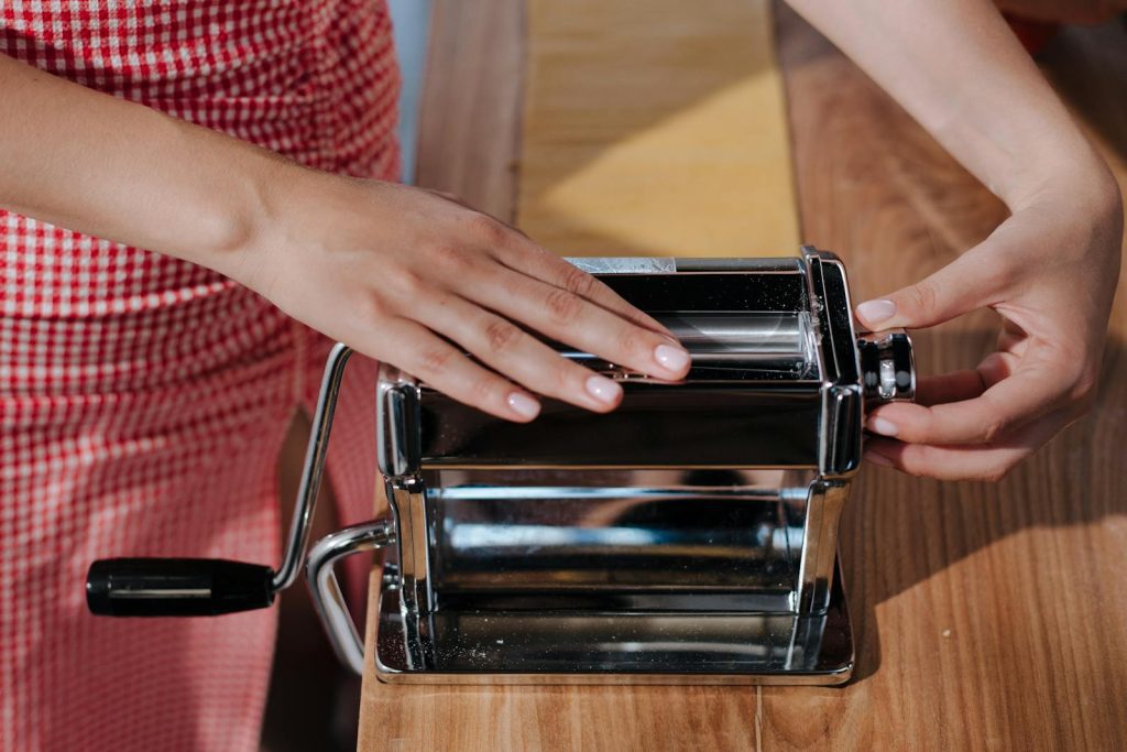 Hands making homemade pasta using a shiny metal pasta maker on a wooden table.